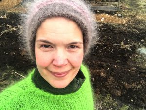 sara står i grön stickad tröja och toppluva i trädgården som är snöfri.