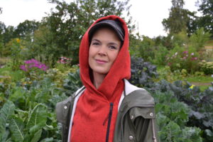 Sara står i röd luvtröja och brun jacka i trädgården och tittar in i kameran.