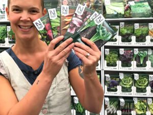 Sara står med fem fröpåsar framför ett ställ med hundratals fröer. Hon ser väldigt glad ut.