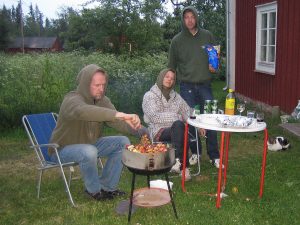 Tre personer grillar i en igenvuxen trädgård framför ett hus.