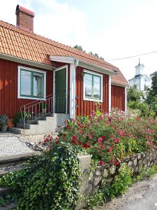 En stenmur och en blomsterrabatt av rosenskäror framför ett rött litet hus.