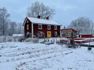Ett rött trähus med växthus och köksträdgård framför, allt insvept i ett vintertäcke.