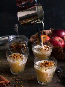 Serveringsglas med fruktkräm och havrecrunch och gräddmjölk, i bakgrunden röda äpplen i brunt papper.