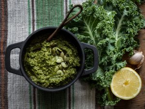 Pesto av grönkål i en svart bytta. Bredvid syns kålblad, citron och vitlöksklyftor.